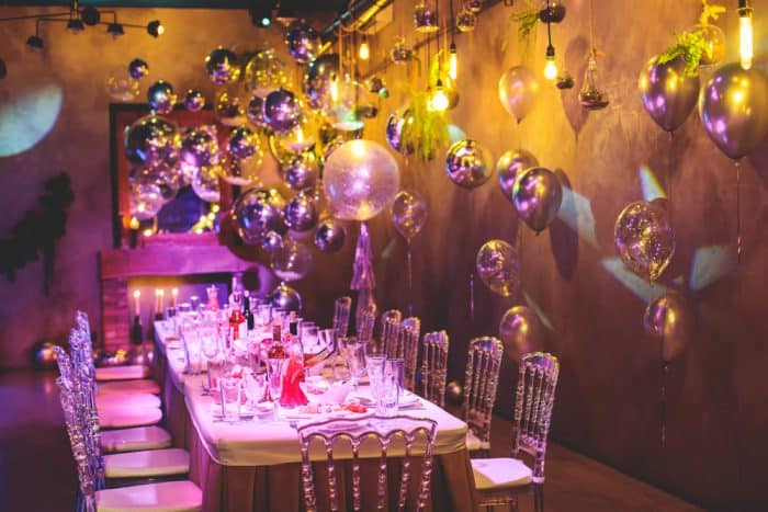 A festive NY dining room with a long table set for guests, decorated with candles, glasses, and plates. Shiny metallic balloons and glowing string lights adorn purple-toned walls, evoking a party hall Brooklyn ambiance.