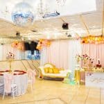 A decorated party hall in Brooklyn, NY features a mirrored wall, chandeliers, and a disco ball. Pink and gold balloon garlands, a white and gold lounge, tables with roses, and a "Melanie" sign create an elegant atmosphere for guests.