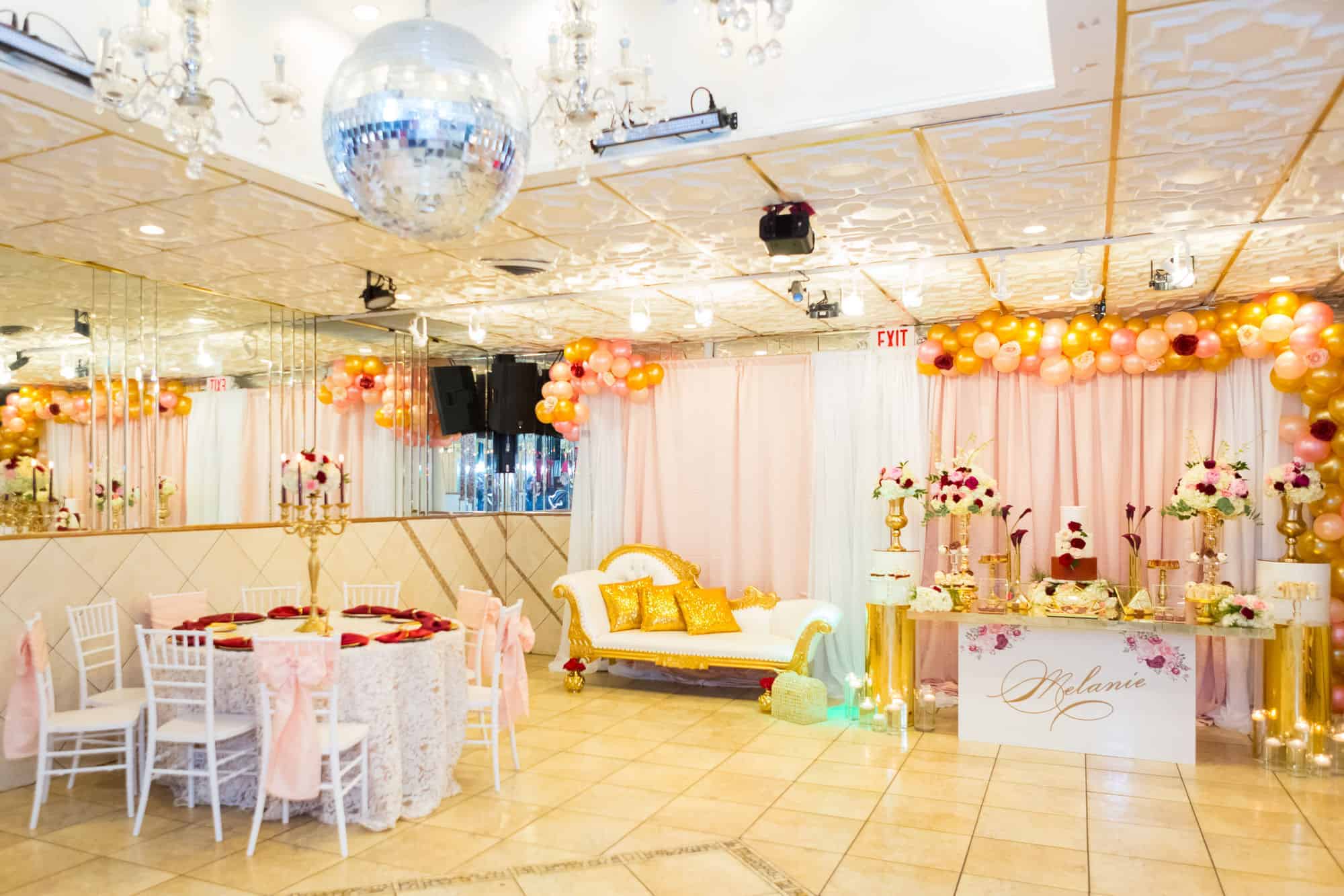 A decorated party hall in Brooklyn, NY features a mirrored wall, chandeliers, and a disco ball. Pink and gold balloon garlands, a white and gold lounge, tables with roses, and a "Melanie" sign create an elegant atmosphere for guests.