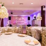 Elegant banquet hall in Brooklyn, NY, decorated for a formal event with round tables covered in white linens, gold chairs, tall floral centerpieces with white and pink flowers, and a stage with draped pink and white fabric in the background.