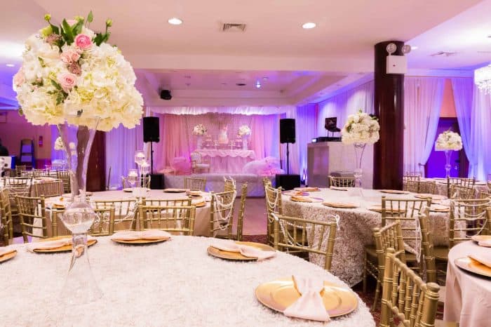 Elegant banquet hall in Brooklyn, NY, decorated for a formal event with round tables covered in white linens, gold chairs, tall floral centerpieces with white and pink flowers, and a stage with draped pink and white fabric in the background.