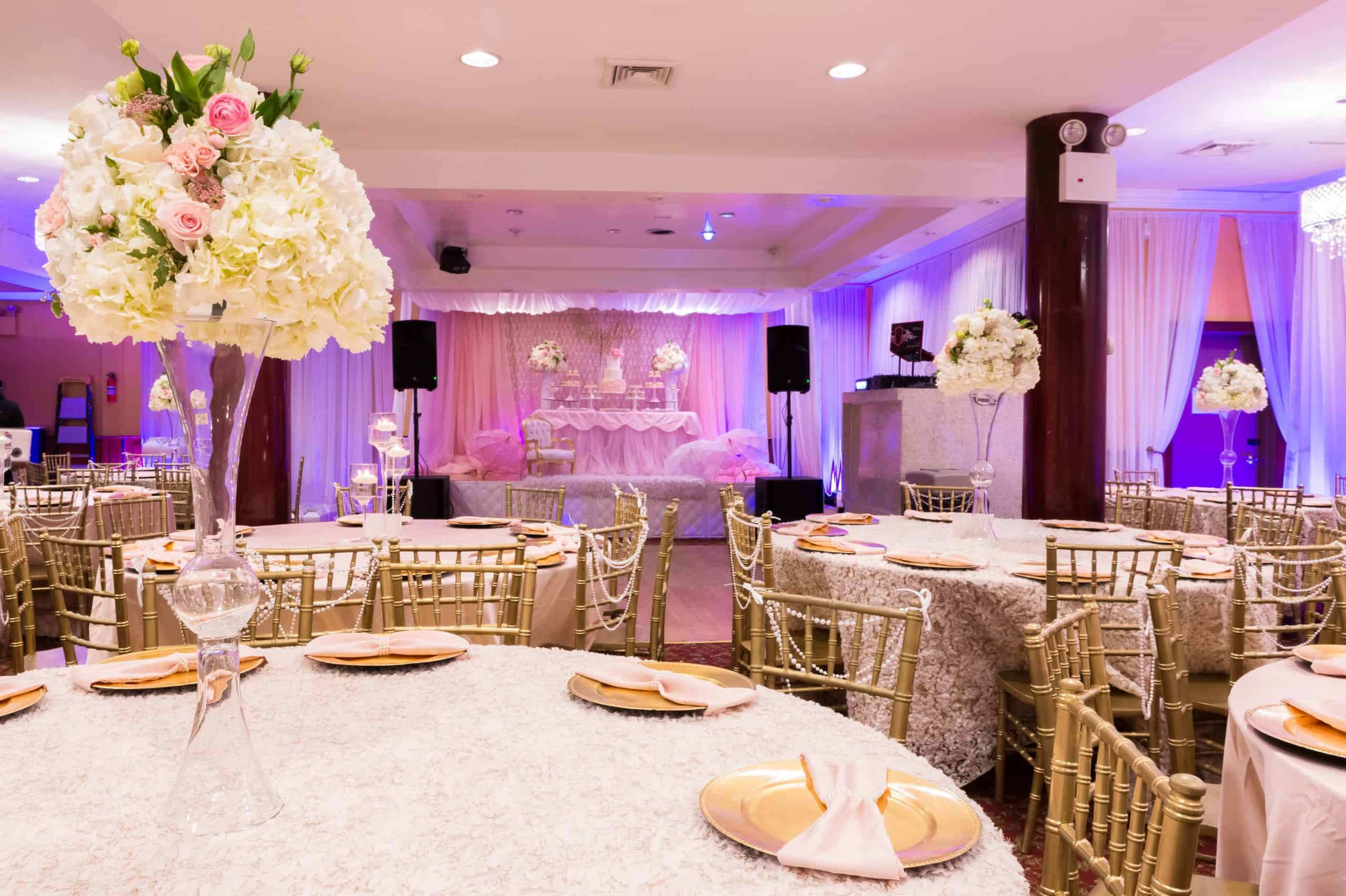 Elegant banquet hall in Brooklyn, NY, decorated for a formal event with round tables covered in white linens, gold chairs, tall floral centerpieces with white and pink flowers, and a stage with draped pink and white fabric in the background.