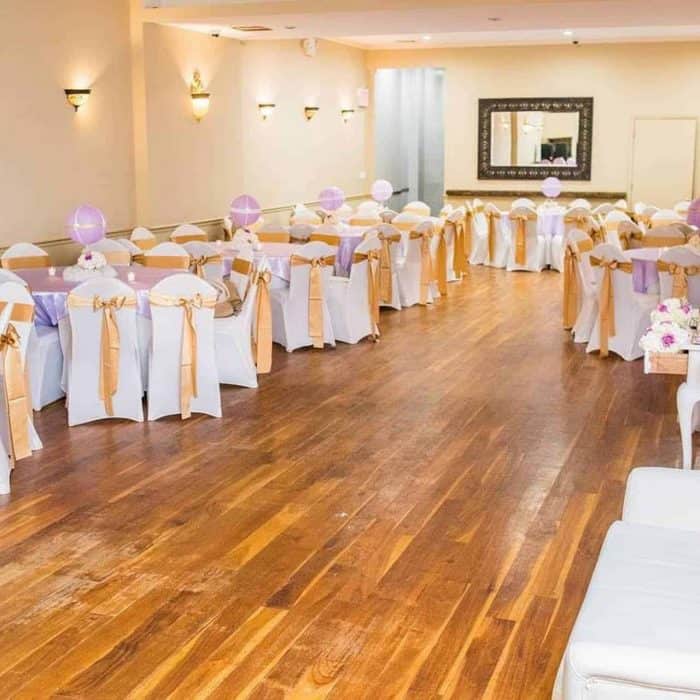 A spacious party hall in Brooklyn, NY with wooden floors, decorated tables and chairs. Chairs have white covers with gold sashes, tables feature white cloths and pastel purple centerpieces, all softly lit for an elegant atmosphere.