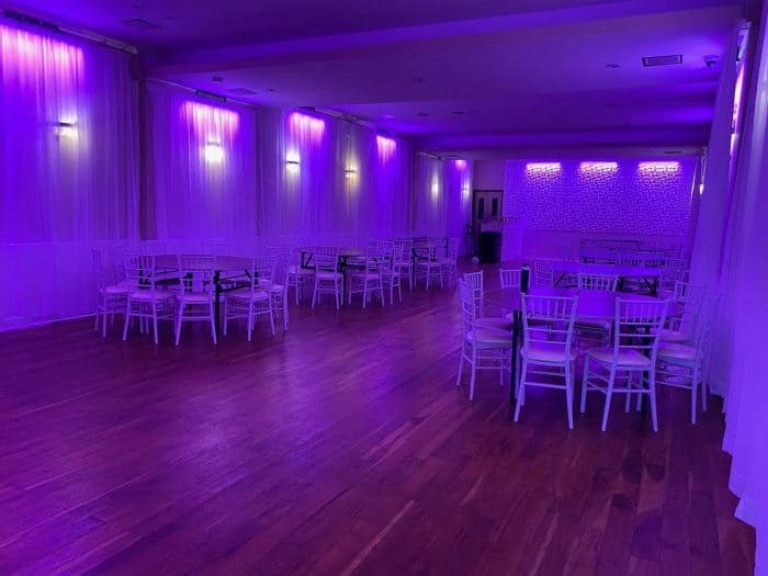 A large, empty party hall in Brooklyn, NY with wooden floors, white chairs and tables, and sheer curtains, all illuminated by vibrant purple LED lighting on the walls and ceiling.