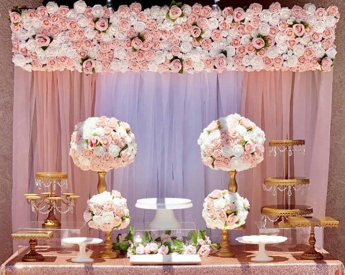 An elegant dessert table in a party hall Brooklyn, NY, decorated with pink and white rose arrangements, gold cake stands, and a pink tulle backdrop bordered with roses—perfectly set for a celebration, though no desserts are yet displayed.