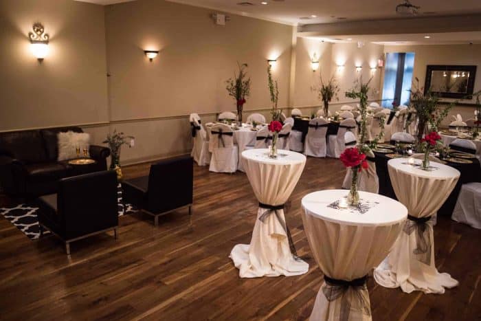 A small party hall in Brooklyn, NY, features high cocktail tables draped in white cloth with vases of red roses. Black chairs and a sofa sit to the side. Wooden floors and soft wall lighting create a warm, elegant ambiance.