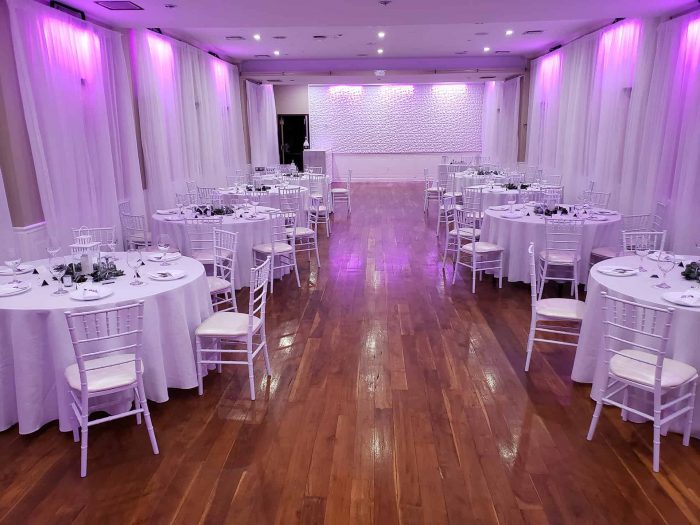 A NY party hall in Brooklyn with polished wooden floors, round tables draped in white cloths, elegant white chairs, and soft purple lighting along the walls—perfectly set up for your next event or celebration.