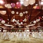 A spacious party hall in Brooklyn, NY, adorned with white and purple paper lanterns hanging from the ceiling, features round tables draped in white cloths and elegant chairs arranged for a formal event.