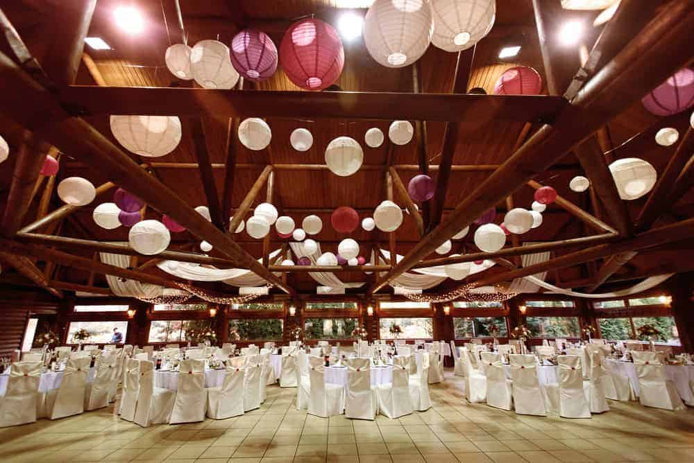 A spacious party hall in Brooklyn, NY, adorned with white and purple paper lanterns hanging from the ceiling, features round tables draped in white cloths and elegant chairs arranged for a formal event.